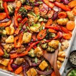Sheet pan filled with cooked chicken pieces, broccoli, red bell peppers, carrots, and red onions, garnished with sesame seeds and sliced green onions, with a wooden spoon.
