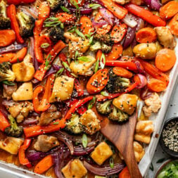 Sheet pan filled with cooked chicken pieces, broccoli, red bell peppers, carrots, and red onions, garnished with sesame seeds and sliced green onions, with a wooden spoon.