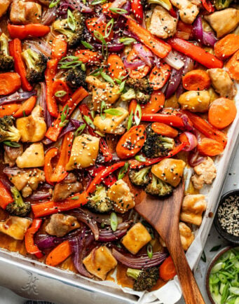 Sheet pan filled with cooked chicken pieces, broccoli, red bell peppers, carrots, and red onions, garnished with sesame seeds and sliced green onions, with a wooden spoon.