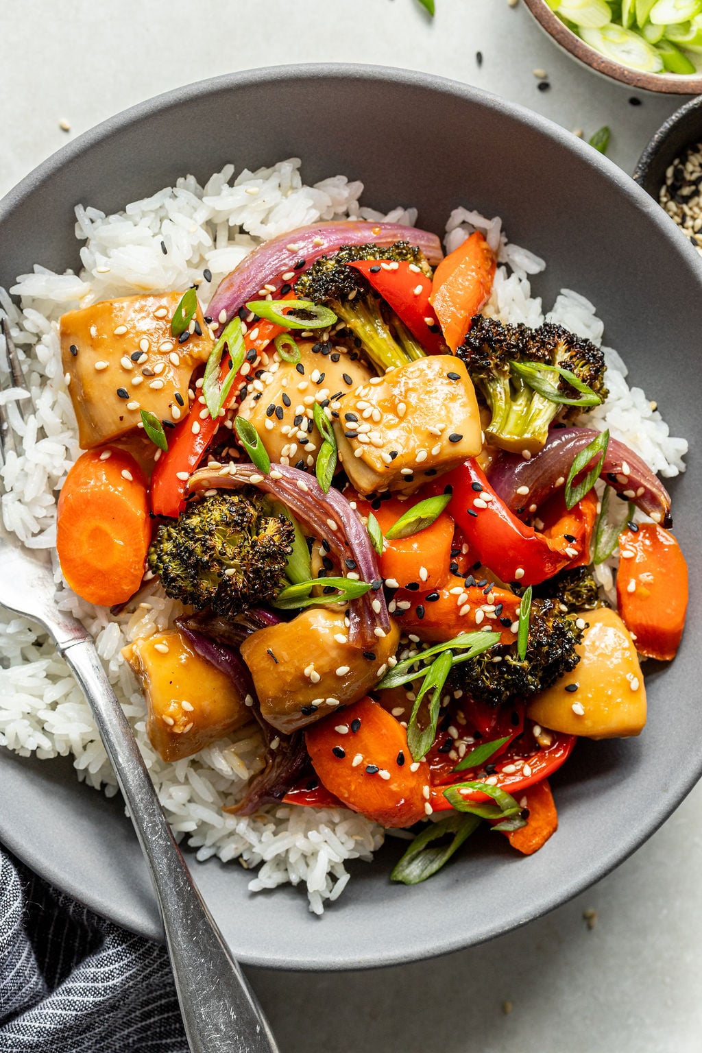 A bowl of white rice topped with stir-fried vegetables, chicken, and sesame seeds, with a fork placed on the side.