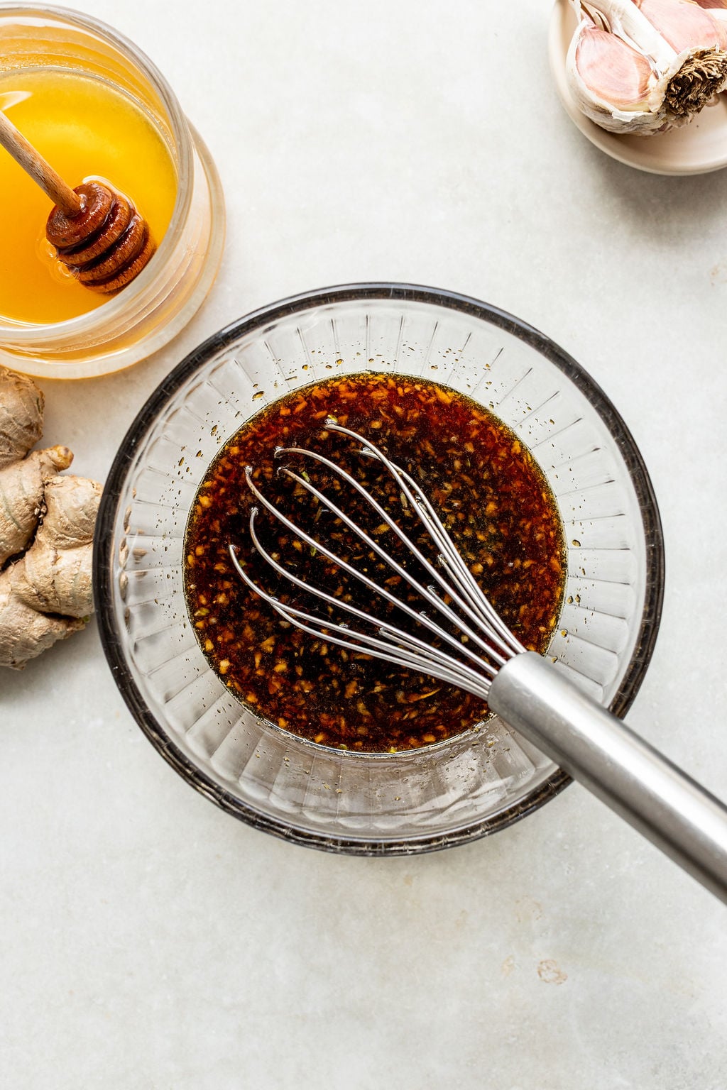 A metal whisk rests in a glass bowl of dark sauce, with honey, ginger root, and garlic displayed nearby on a light surface.
