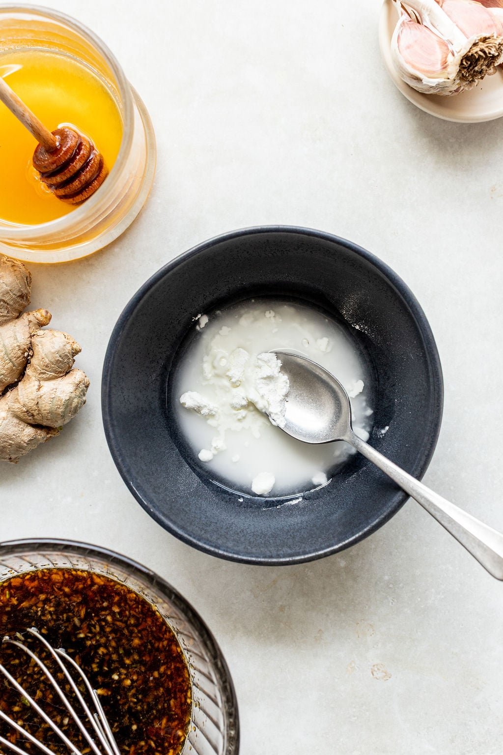 A black bowl with a spoon holding a white mixture, surrounded by honey, ginger, garlic, and a bowl of dark sauce on a light surface.