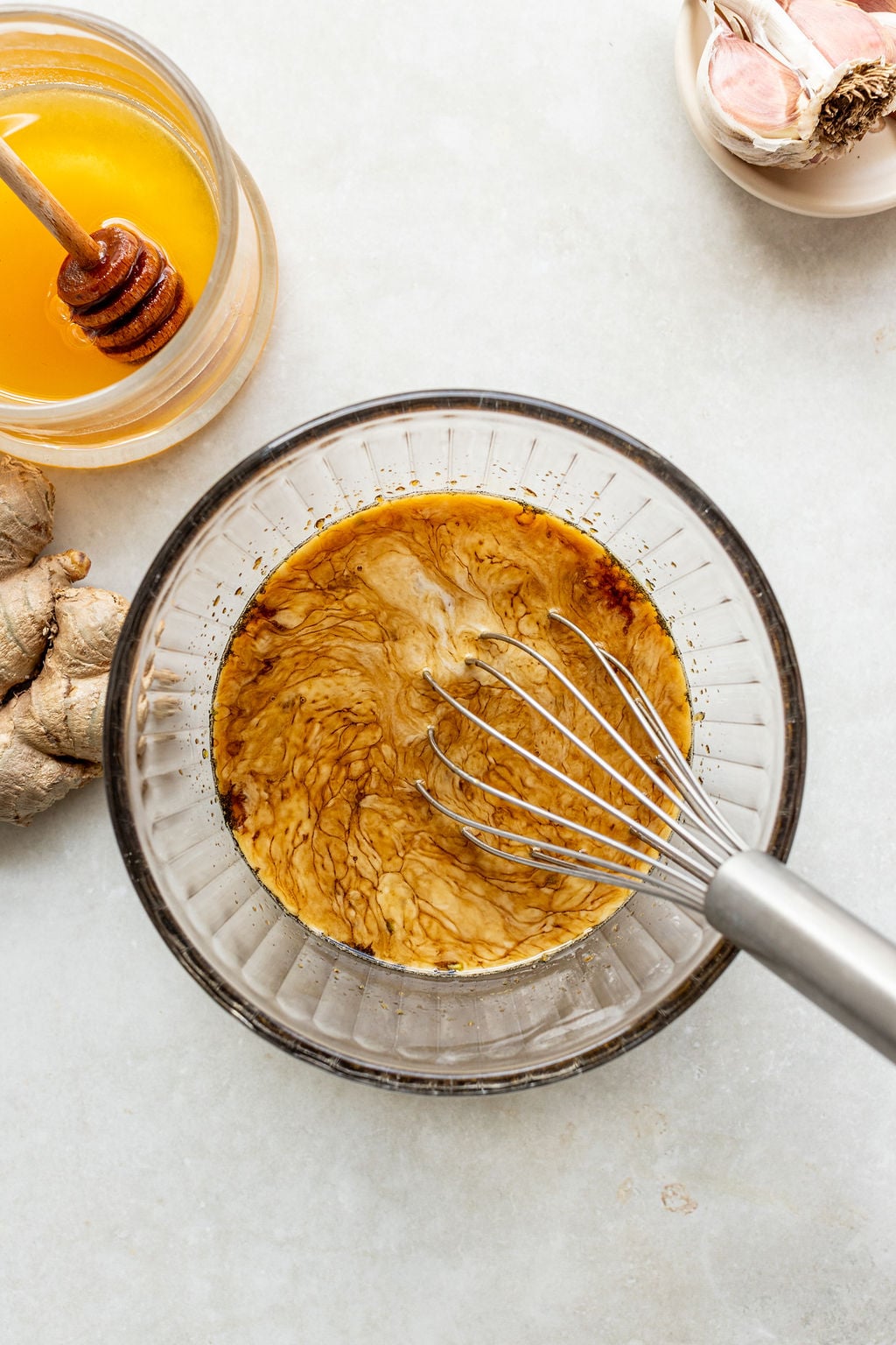 A metal whisk in a glass bowl mixing a brown liquid; ginger, honey, and garlic are nearby on a light surface.
