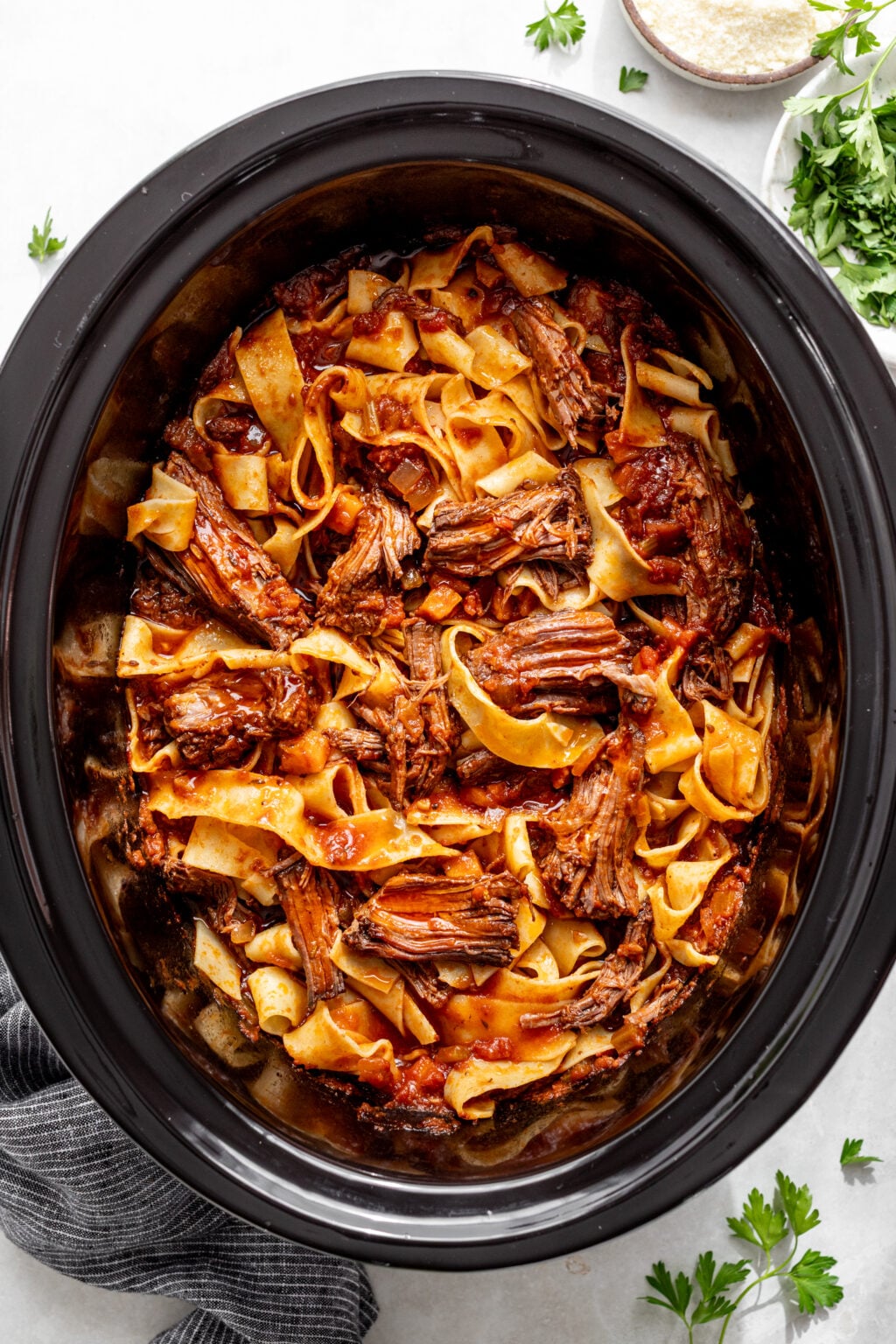A slow cooker filled with shredded beef, wide pasta noodles, and tomato sauce, surrounded by a napkin, fresh parsley, and a bowl of grated cheese.