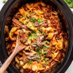 A slow cooker filled with pasta, shredded meat in tomato sauce, topped with chopped parsley and grated cheese, with a wooden spoon resting inside. Parsley and cheese bowls are nearby.