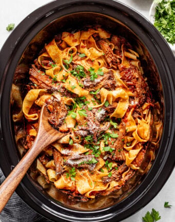 A slow cooker filled with pasta, shredded meat in tomato sauce, topped with chopped parsley and grated cheese, with a wooden spoon resting inside. Parsley and cheese bowls are nearby.