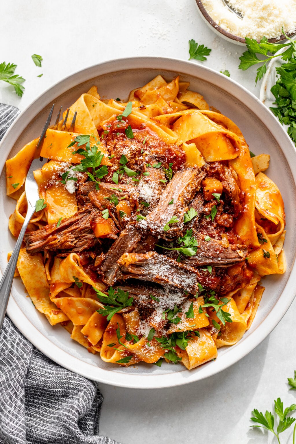 A plate of pappardelle pasta topped with shredded beef ragu, tomato sauce, grated cheese, and fresh parsley, with a fork on the side.