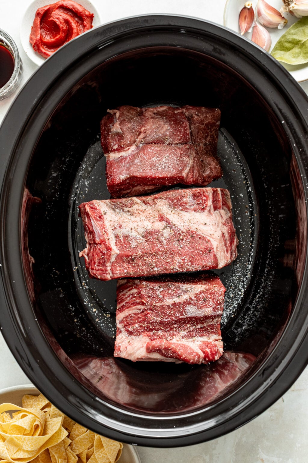 Three raw beef short ribs seasoned with salt and pepper are placed in a black slow cooker. Surrounding the cooker are bowls with pasta, tomato paste, garlic, bay leaves, and red wine.