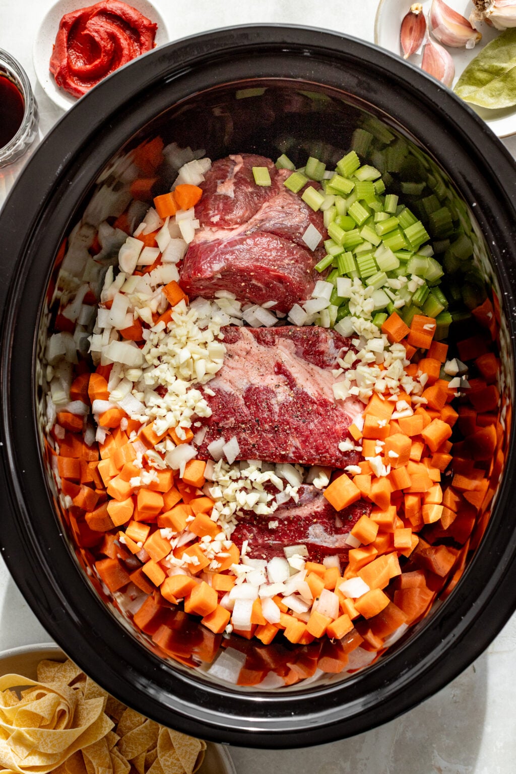 Raw beef, chopped carrots, celery, onions, and garlic are arranged in a slow cooker. Surrounding bowls hold tomato paste, red wine, bay leaves, pasta, and garlic cloves.