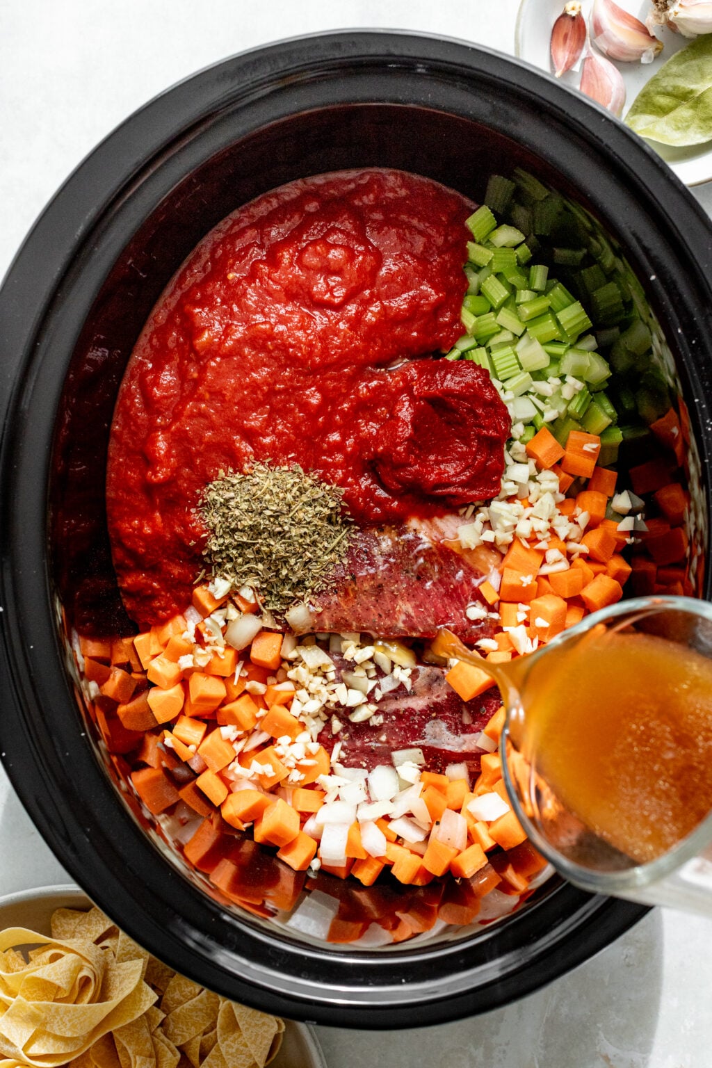 A slow cooker filled with diced carrots, celery, onions, garlic, ground meat, tomato sauce, tomato paste, herbs, and broth being poured in, ready to be cooked.