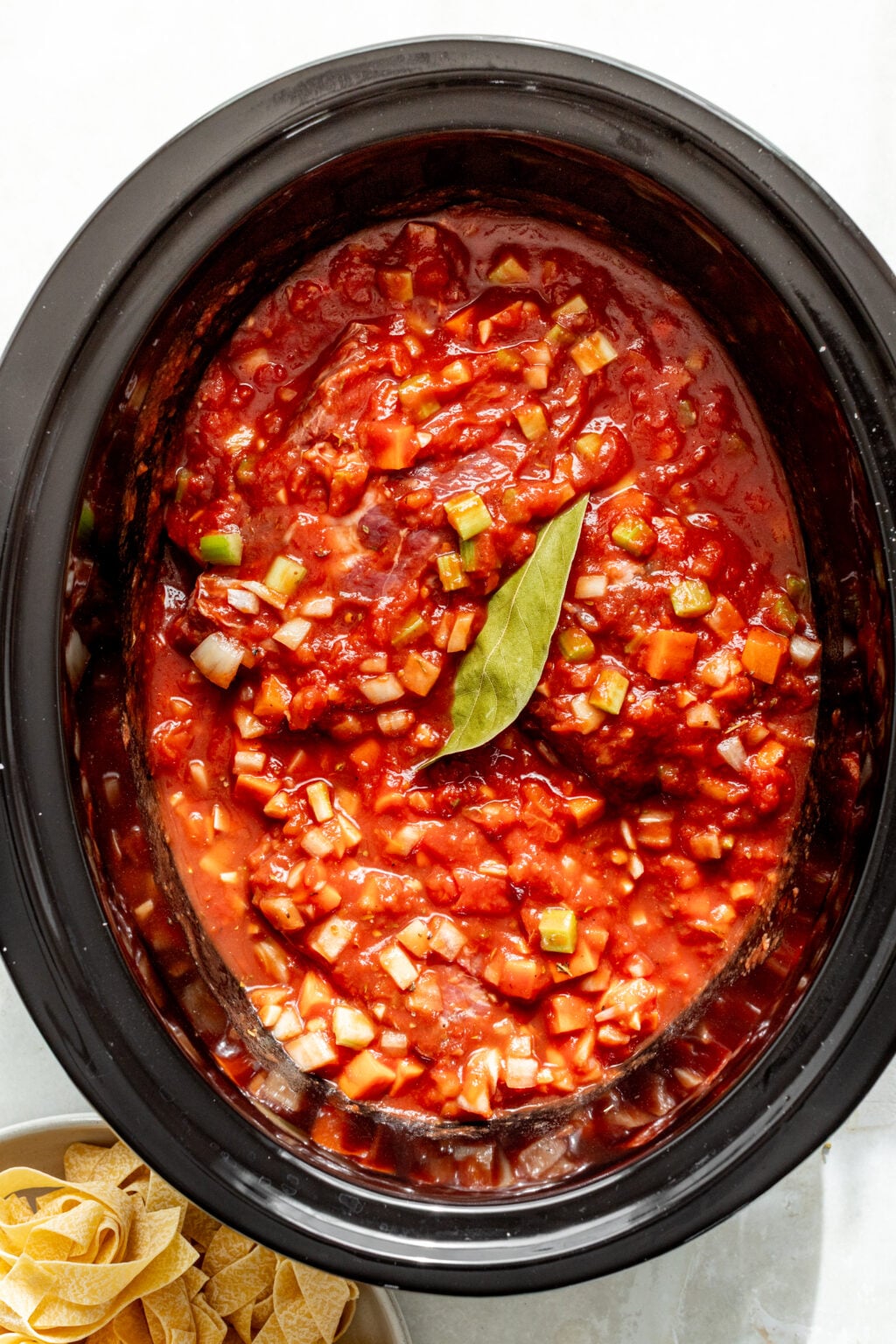 A slow cooker filled with tomato-based sauce containing diced vegetables and a bay leaf, with a bowl of pasta beside it.