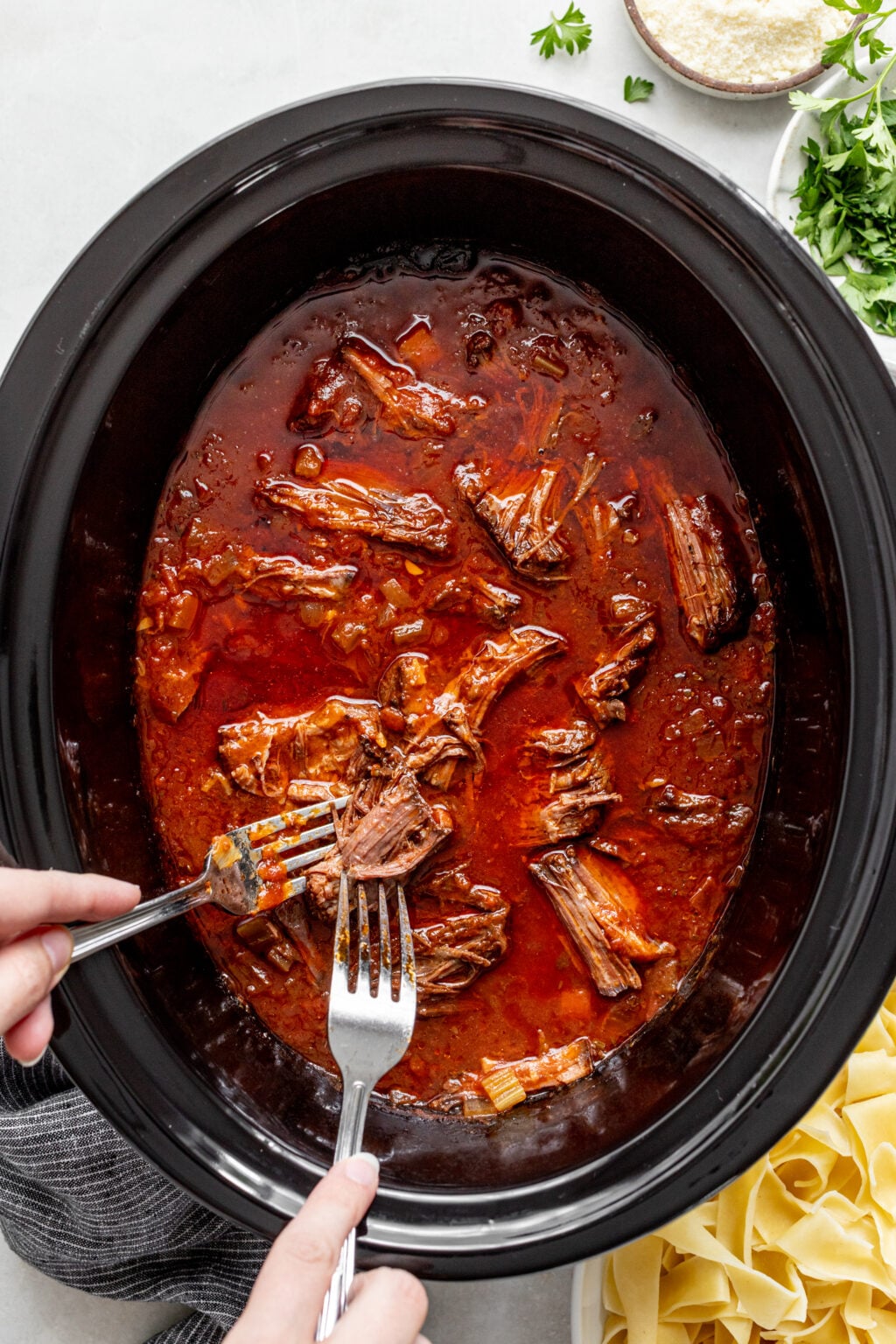 Hands shredding cooked beef with two forks in a slow cooker filled with red sauce, with parsley, parmesan, and pasta visible on the side.