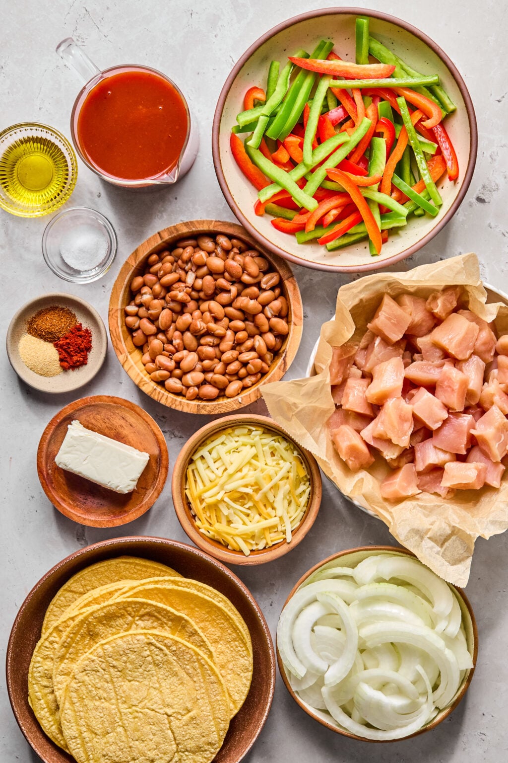 Assorted ingredients for a fajita dish, including raw chicken, sliced onions, bell peppers, pinto beans, cheese, cream cheese, tortillas, sauce, oil, and spices on a countertop.