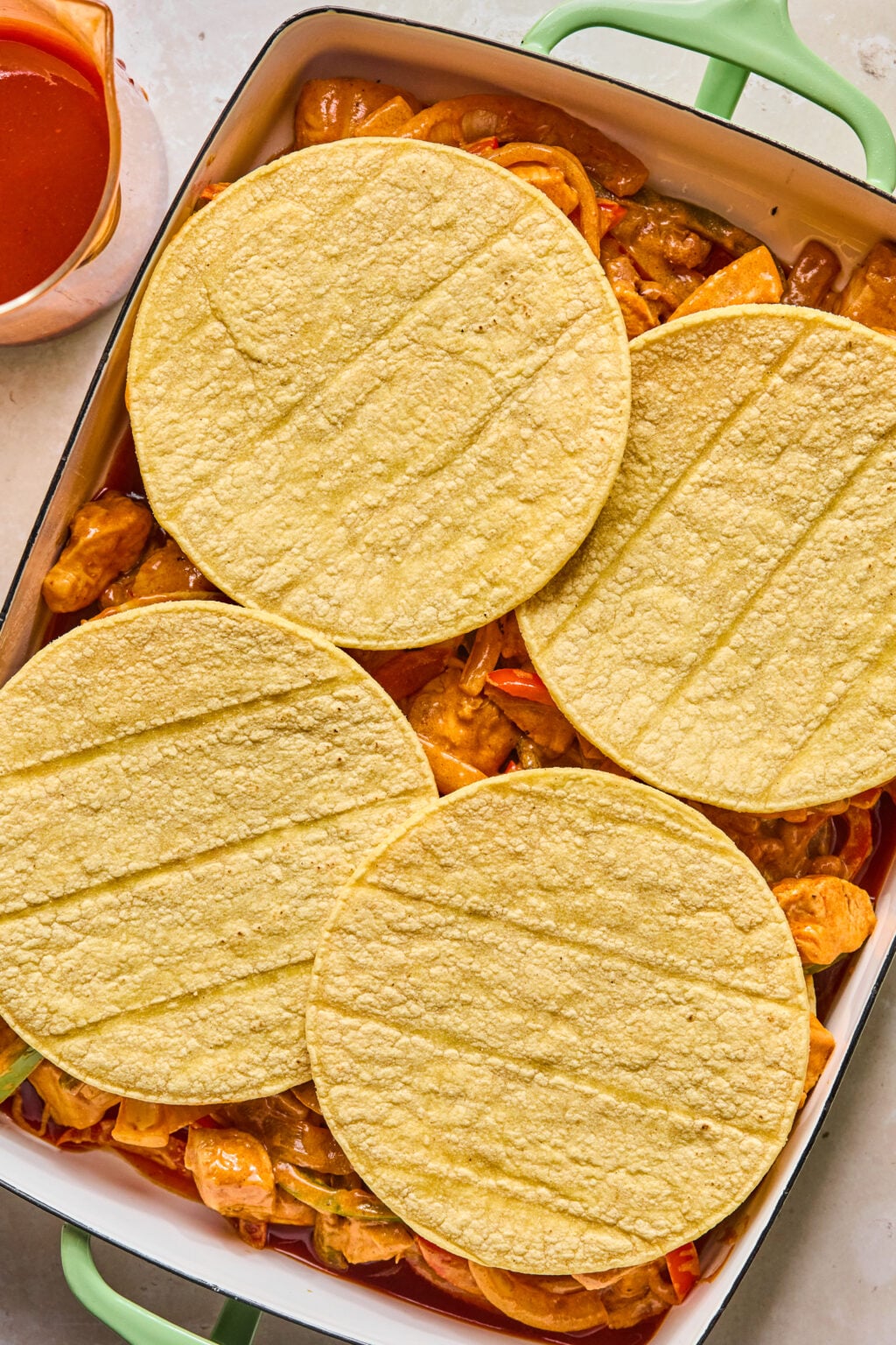 Four corn tortillas are layered over a mixture of vegetables and sauce in a green baking dish, with a cup of red sauce nearby.