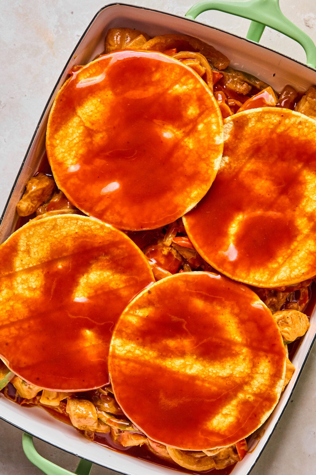 Four corn tortillas topped with red enchilada sauce are layered over a mixture of cooked ingredients in a green-handled baking dish.