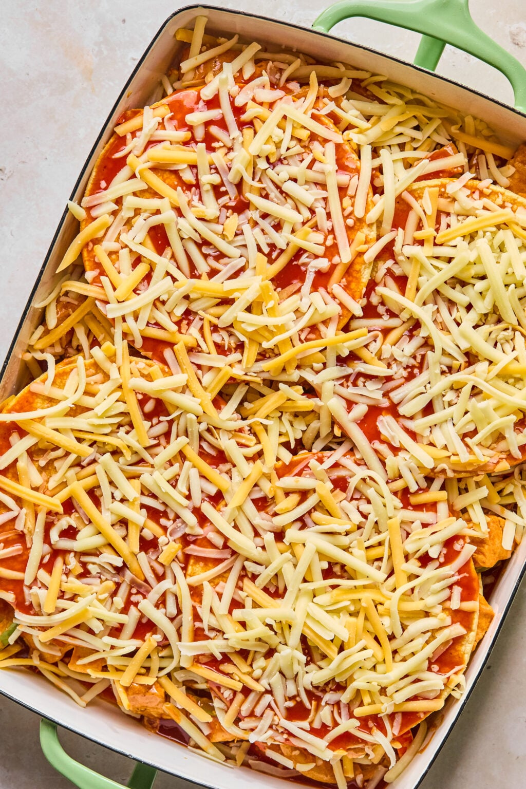 A rectangular baking dish filled with tortillas, red sauce, and shredded cheese, ready to be baked.