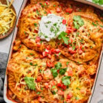A casserole dish filled with cheesy enchiladas, topped with sour cream, chopped tomatoes, and cilantro, sits on a table with shredded cheese and a glass of water nearby.