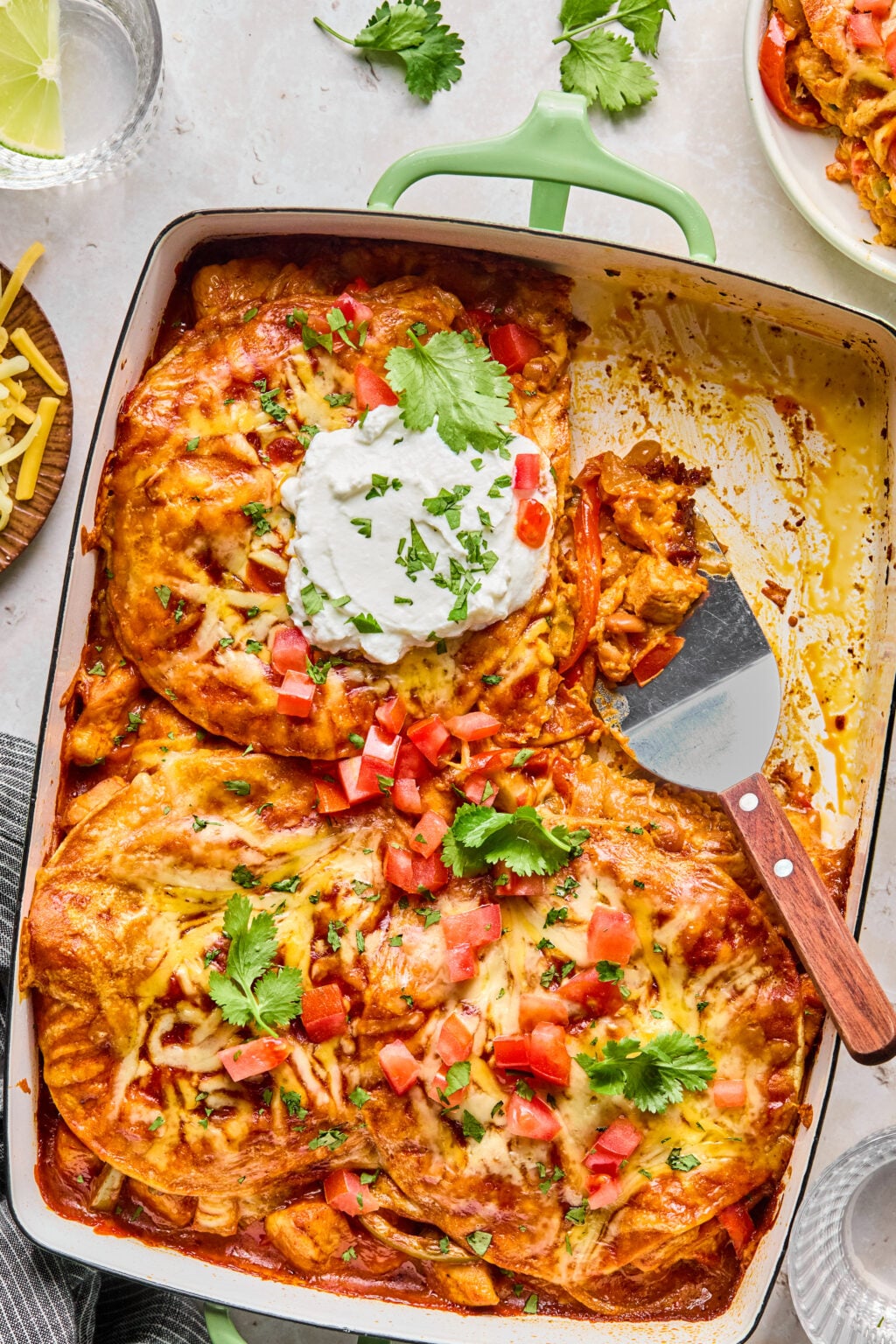 A casserole dish filled with baked enchiladas topped with melted cheese, diced tomatoes, cilantro, and a dollop of sour cream, with a serving spatula on the side.