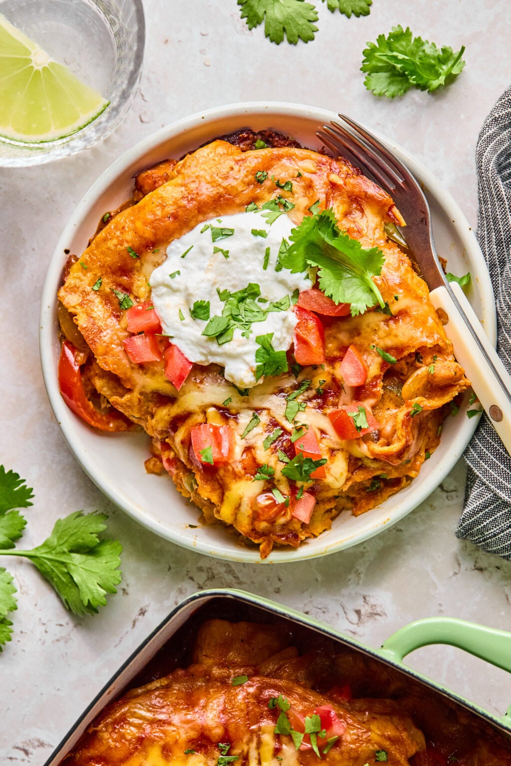 A plate of cheesy enchiladas topped with sour cream, chopped tomatoes, and cilantro, with a fork and knife beside it. A glass of water with lime and cilantro garnish are nearby.