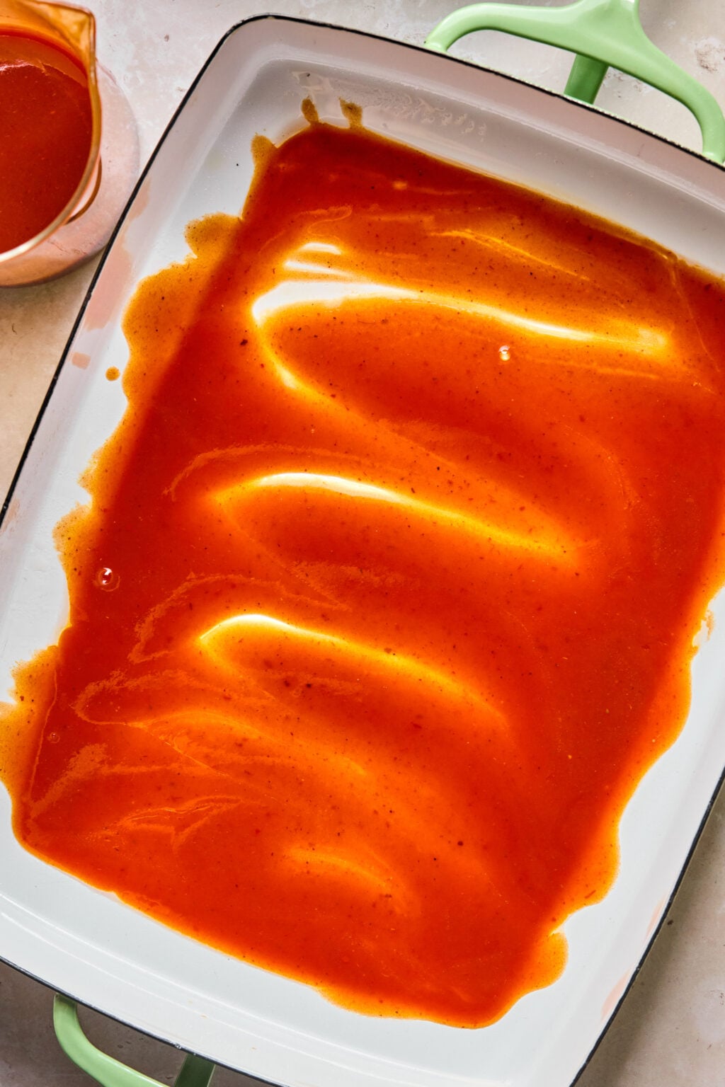 A baking dish with a layer of red enchilada sauce spread across the bottom, with a small pitcher of sauce in the corner.