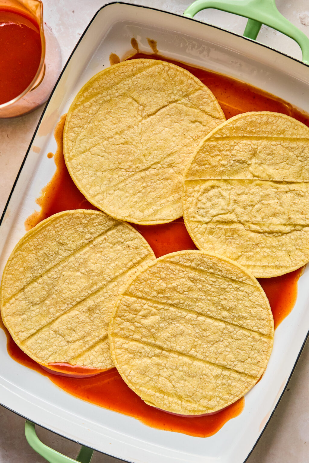 Four corn tortillas are laid flat in a white baking dish with red enchilada sauce partially covering the bottom. A small pitcher with more sauce is nearby.