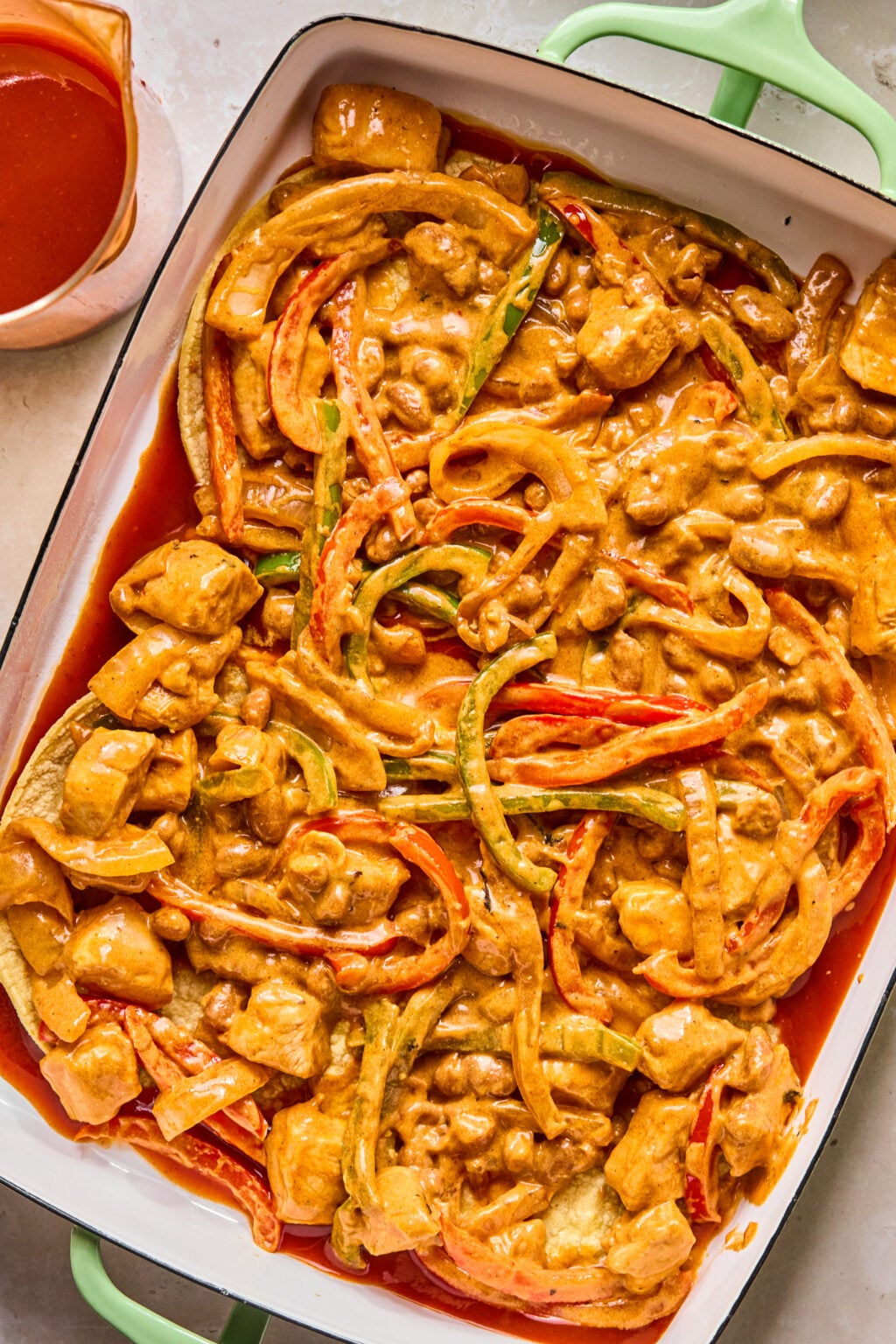 A casserole dish filled with creamy, orange-colored curry containing sliced bell peppers, chunks of chicken, and sauce, with extra sauce in a cup nearby.
