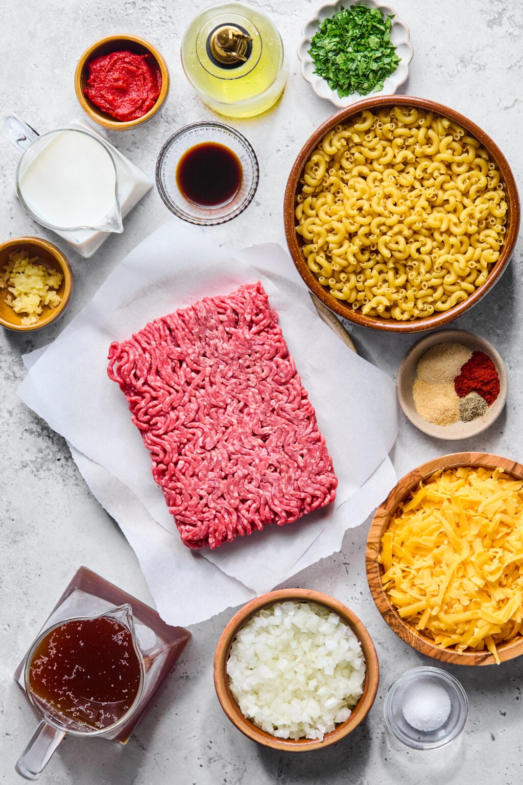 Ingredients for a pasta dish arranged on a counter, including raw ground beef, elbow macaroni, shredded cheese, chopped onion, broth, milk, spices, sauce, and herbs in bowls.