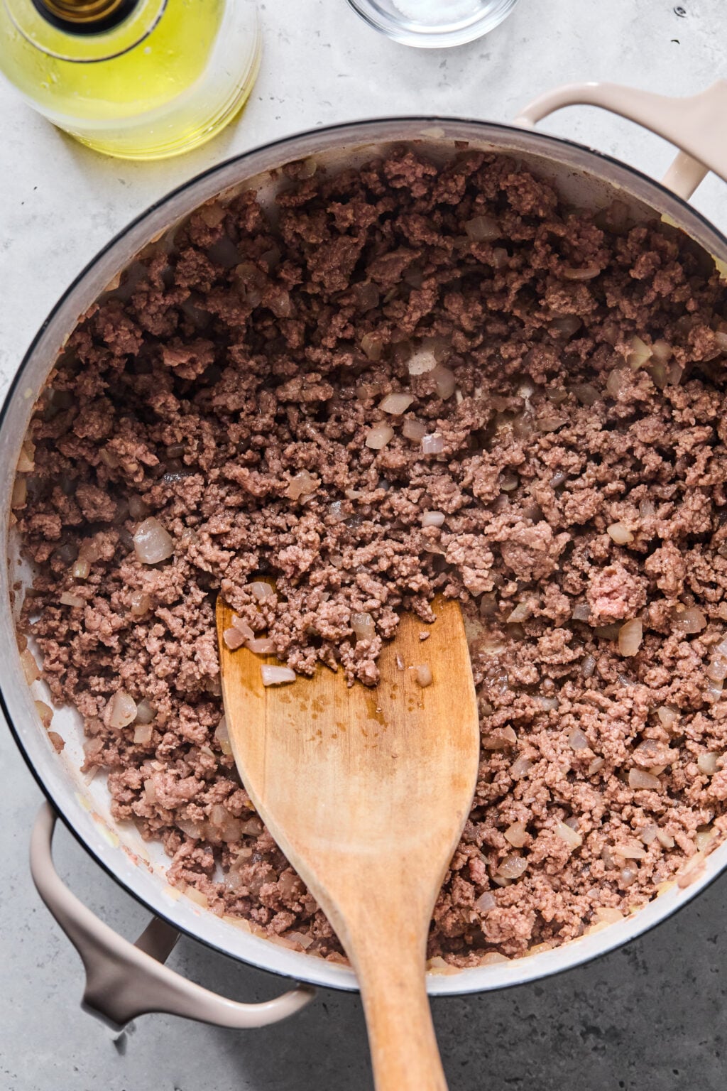 Ground beef and diced onions being cooked in a large pan, stirred with a wooden spoon. A bottle of oil and a glass are visible nearby on the countertop.