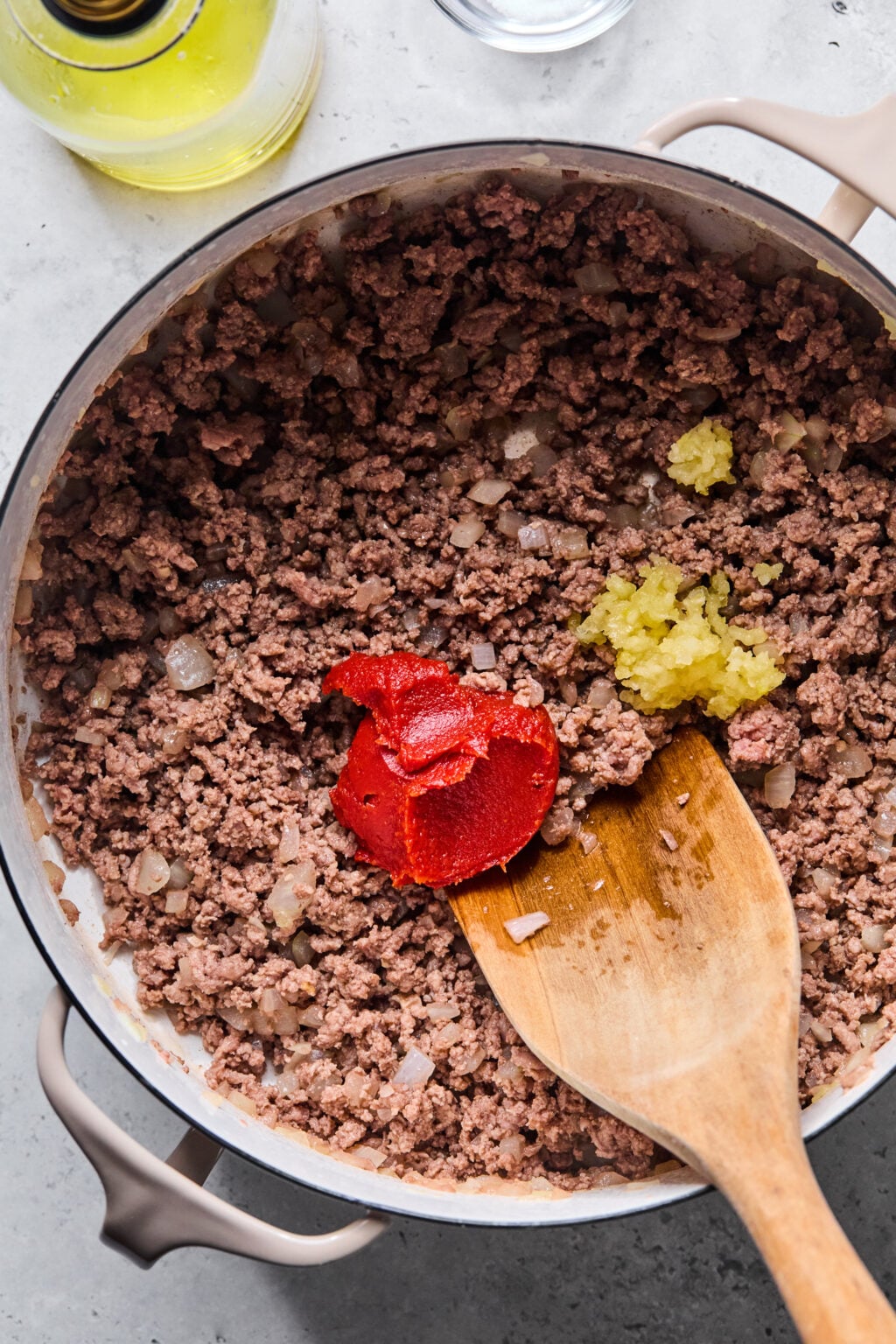 Ground beef and diced onions cooking in a pot with a wooden spoon, topped with tomato paste and minced garlic, on a gray countertop.