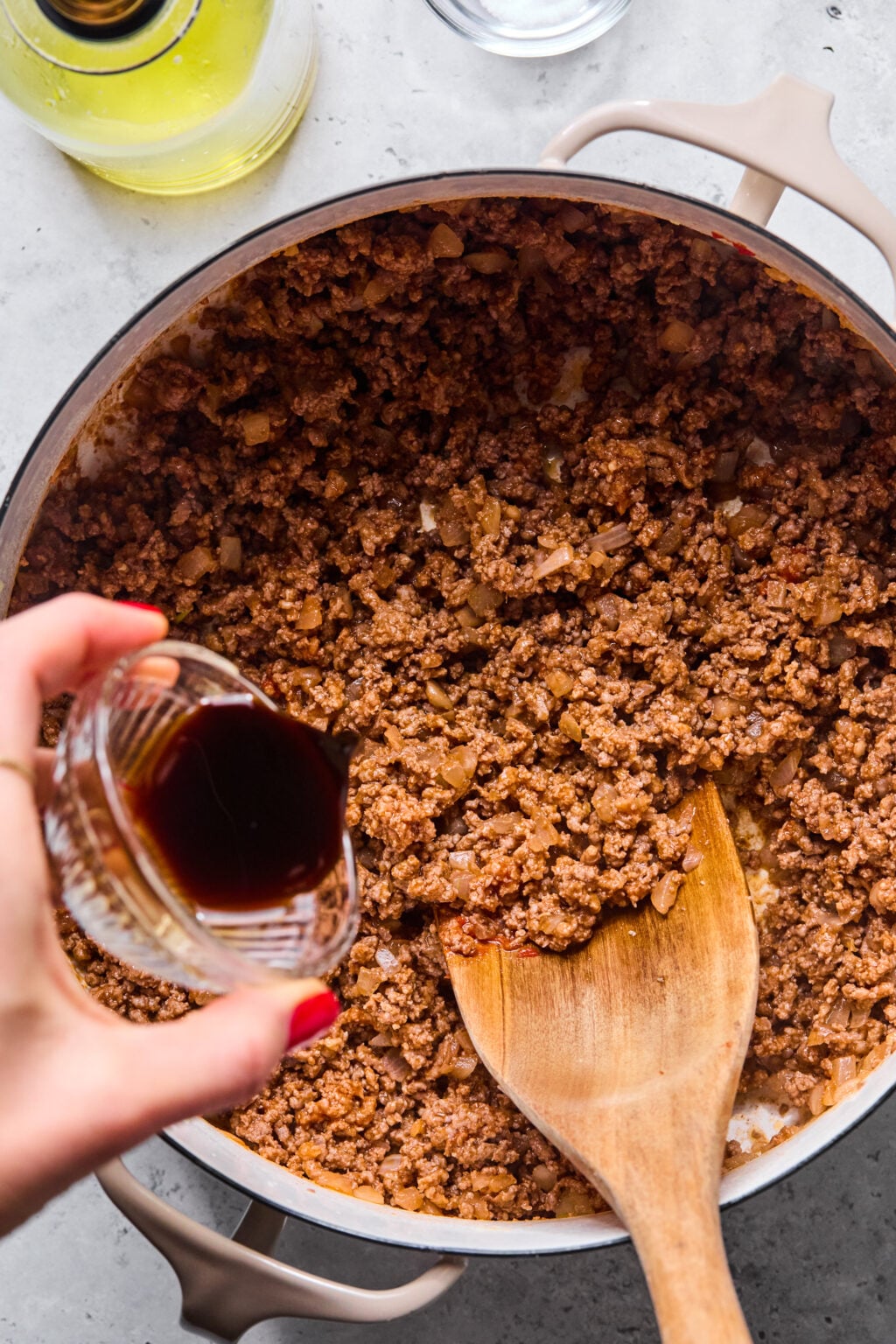Ground beef and onions are being cooked in a Dutch oven, while a hand pours a dark sauce from a small glass bowl into the mixture with a wooden spoon.