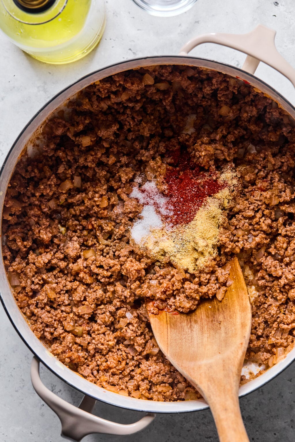 Ground beef cooking in a pot with visible seasonings on top, being stirred with a wooden spoon; a bottle of oil is nearby.