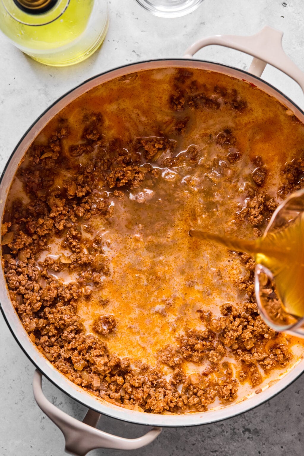 Ground beef simmering in a pot, with liquid being poured in from a measuring cup, next to a bottle of oil on a gray countertop.