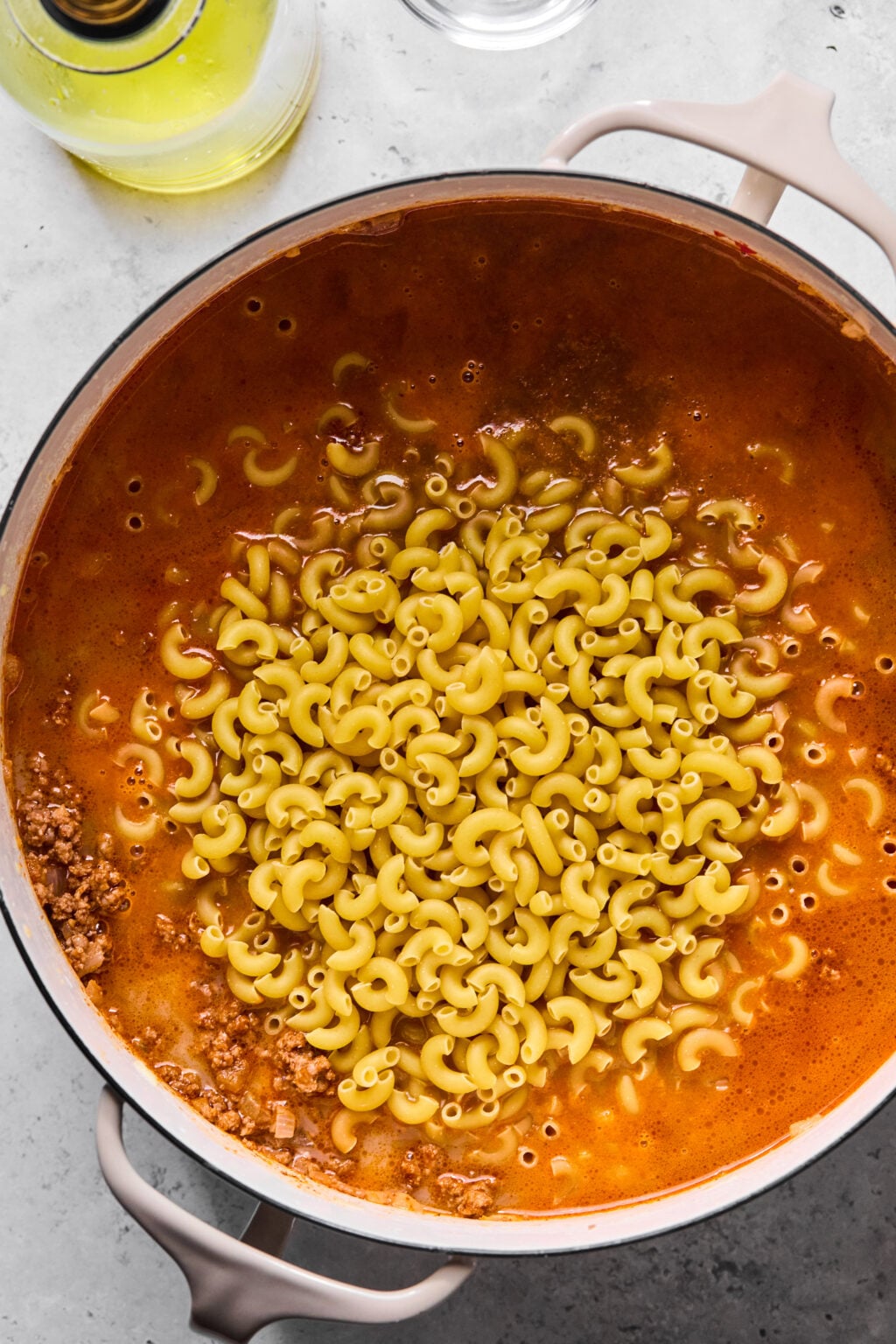 A pot of uncooked elbow macaroni added to a simmering mixture of ground meat and tomato-based sauce on a stovetop.