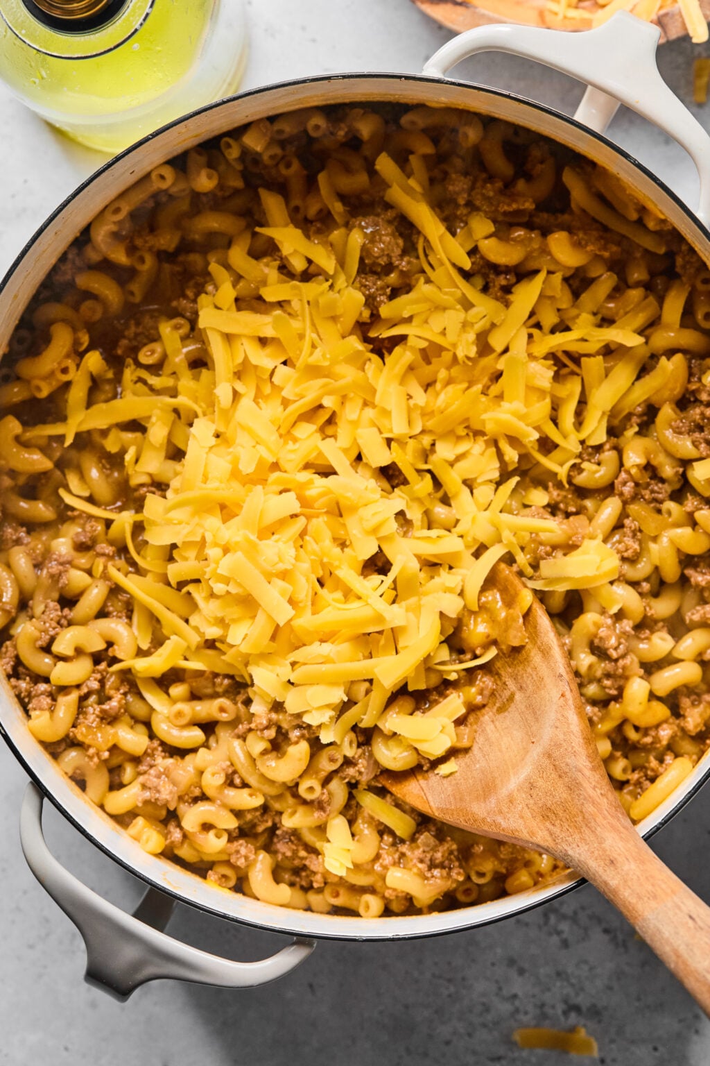 A pot of macaroni mixed with ground meat and topped with shredded cheddar cheese, being stirred with a wooden spoon.