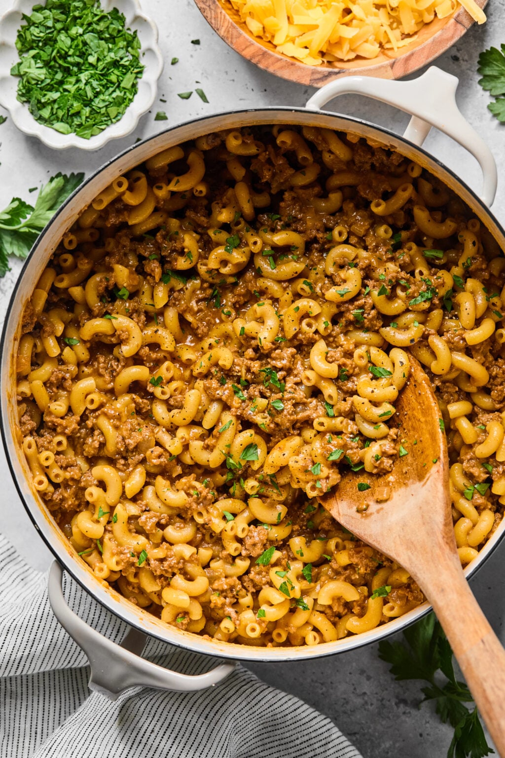 A pot of macaroni with ground meat sauce, garnished with chopped herbs, with a wooden spoon resting in the dish. Bowls of chopped herbs and shredded cheese are nearby.