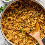 A pot of macaroni with ground meat sauce, garnished with chopped herbs, with a wooden spoon resting in the dish. Bowls of chopped herbs and shredded cheese are nearby.