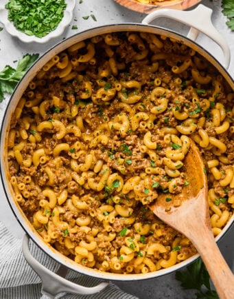 A pot of macaroni with ground meat sauce, garnished with chopped herbs, with a wooden spoon resting in the dish. Bowls of chopped herbs and shredded cheese are nearby.