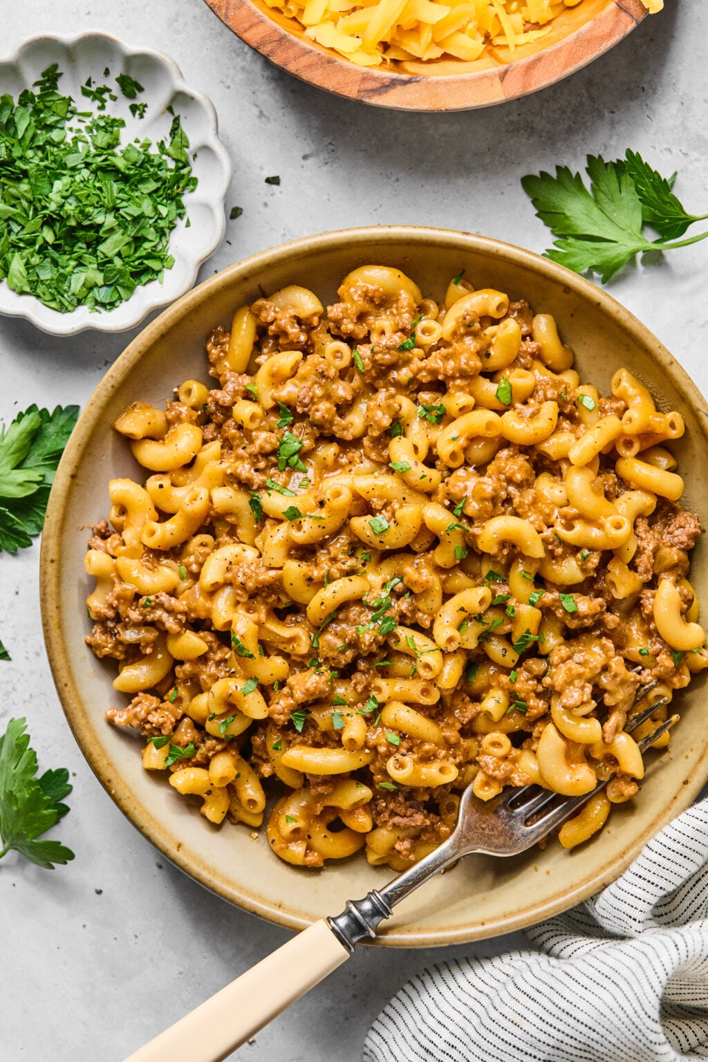 A bowl of macaroni mixed with ground beef and herbs, garnished with chopped parsley, with a fork and small bowls of grated cheese and parsley on the side.