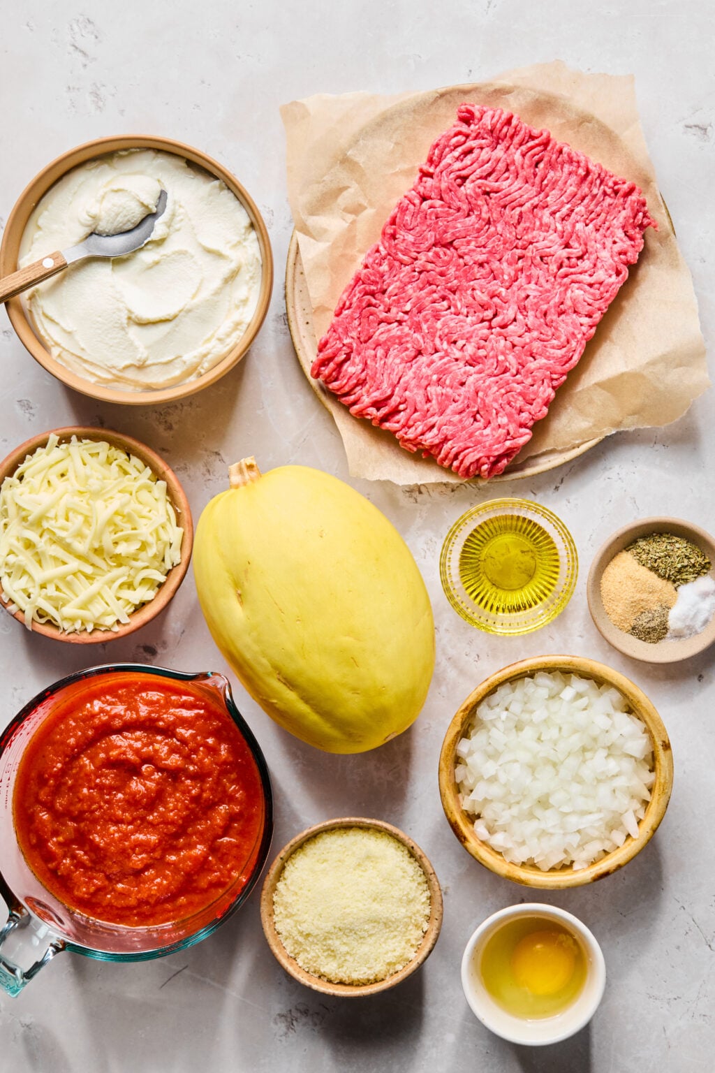 A flat lay of ground beef, spaghetti squash, ricotta cheese, shredded mozzarella, marinara sauce, chopped onion, grated parmesan, an egg, olive oil, and assorted spices on a light surface.