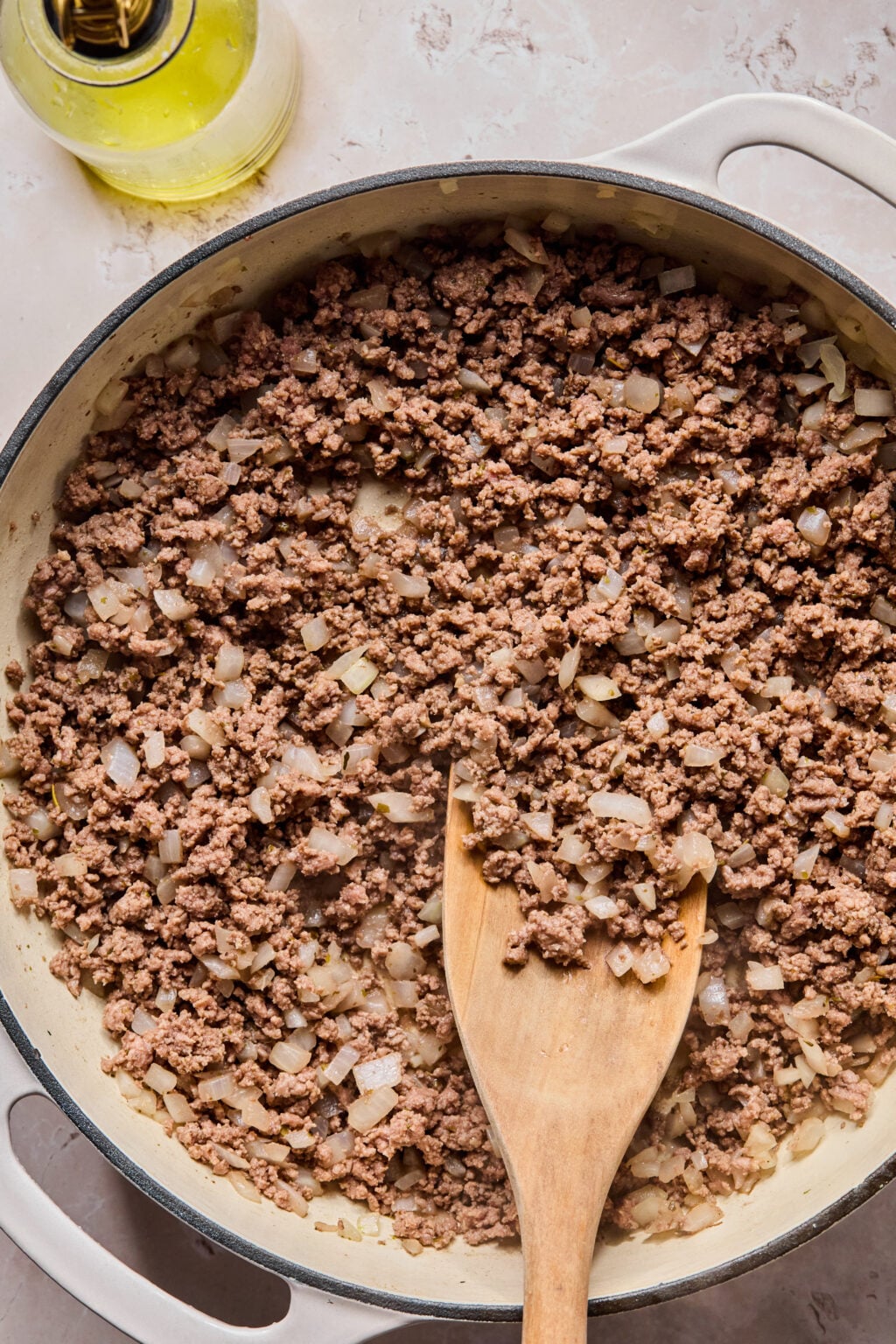 Ground beef and diced onions are being cooked in a white skillet, stirred with a wooden spatula; a bottle of oil is visible in the corner.