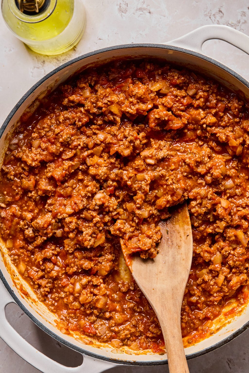 A pot of ground meat and tomato sauce being stirred with a wooden spoon, with a bottle of olive oil nearby.