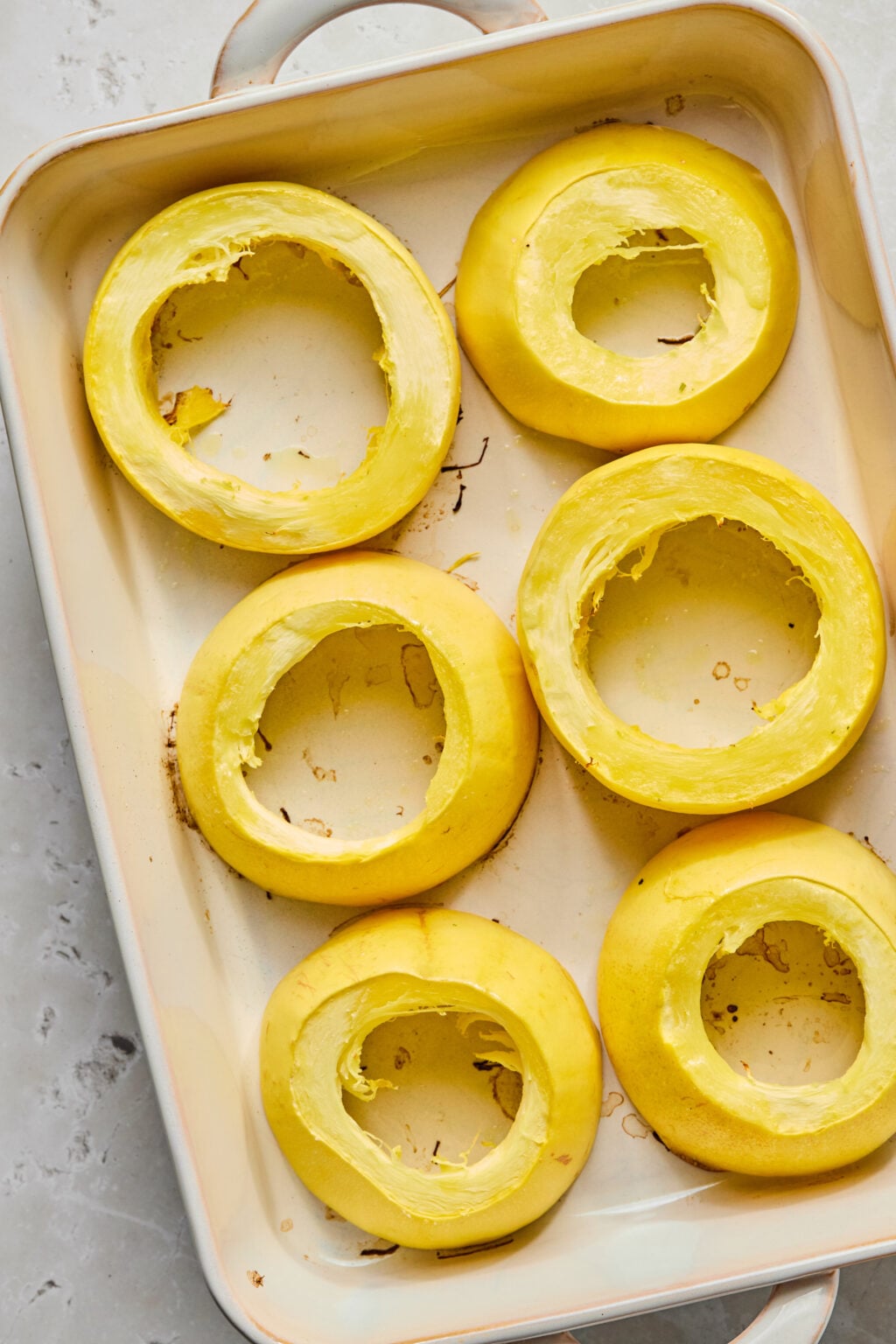 Six hollowed-out yellow squash rings arranged in a single layer in a white baking dish on a light-colored surface.