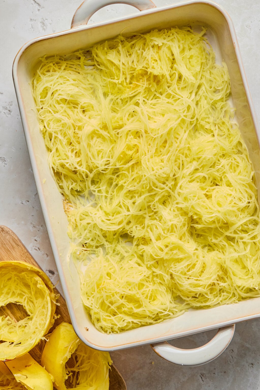 Cooked spaghetti squash strands in a white baking dish, with empty squash shells on a wooden cutting board beside it.