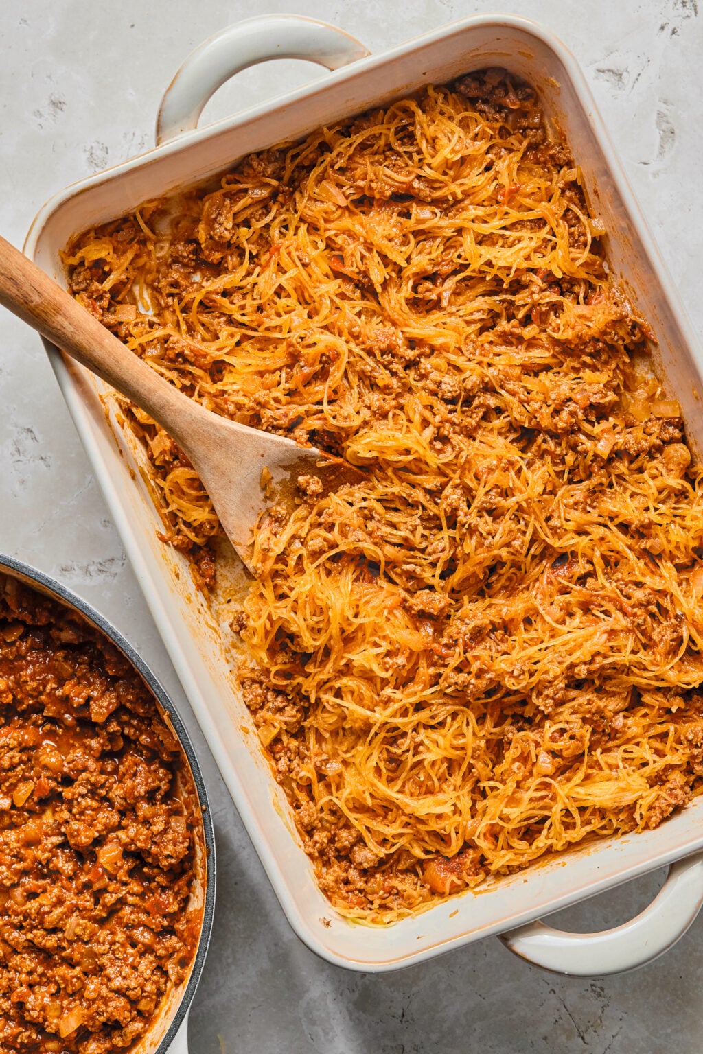 A baking dish filled with spaghetti squash and meat sauce, partially stirred with a wooden spoon, next to a pan containing more meat sauce.