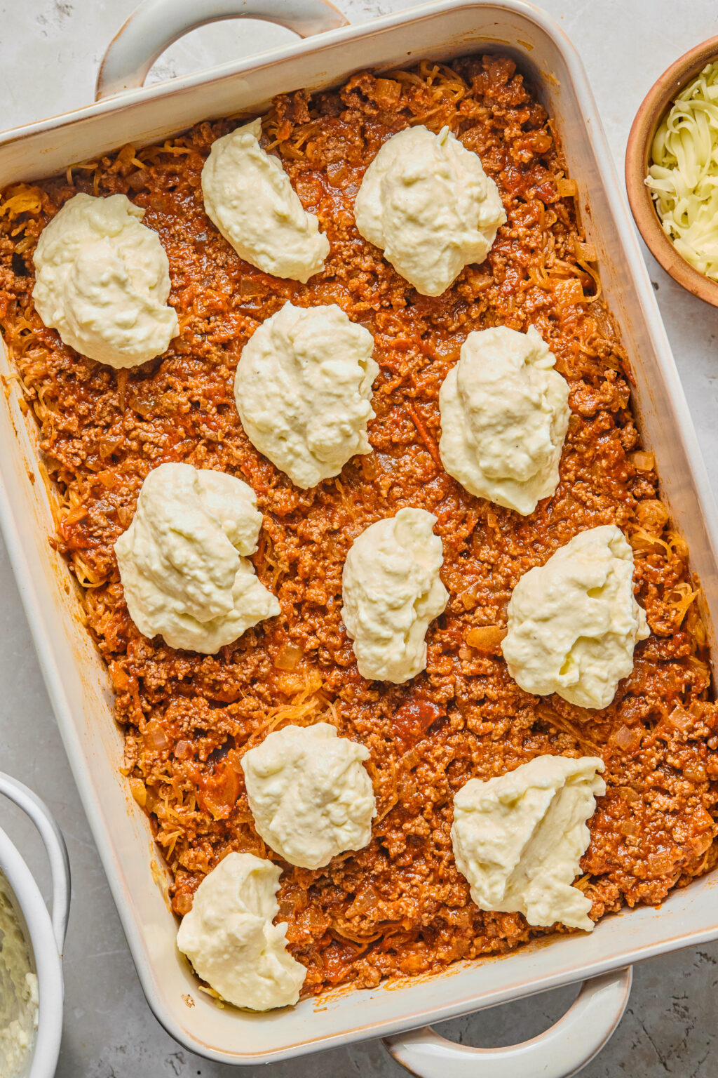 A casserole dish filled with a layer of meat sauce topped with dollops of ricotta cheese mixture, ready to be spread. A bowl of shredded cheese is visible nearby.