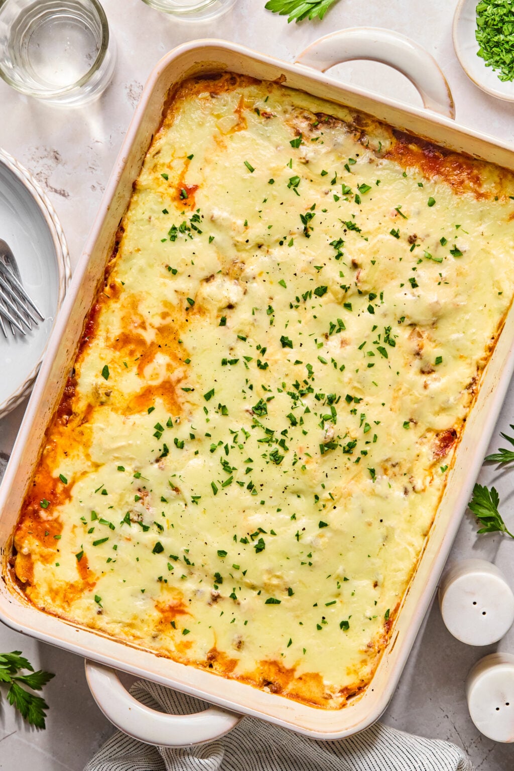 A baked casserole dish topped with melted cheese and chopped herbs, surrounded by plates, utensils, and small bowls of ingredients on a white surface.