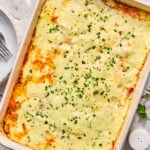 A baked casserole dish topped with melted cheese and chopped herbs, surrounded by plates, utensils, and small bowls of ingredients on a white surface.