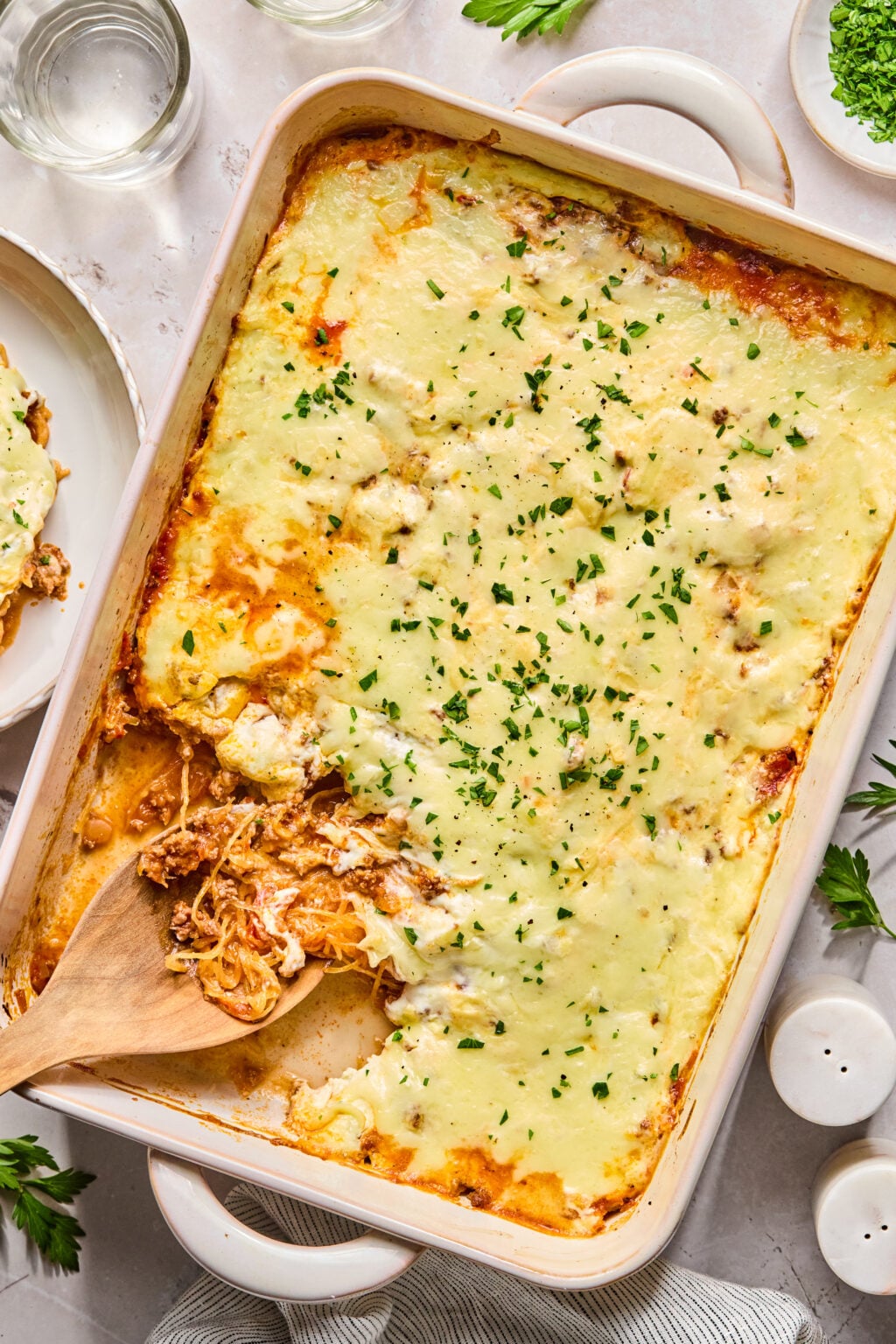 A baked casserole in a rectangular dish topped with melted cheese and chopped parsley, with one serving removed and a wooden spoon resting inside.