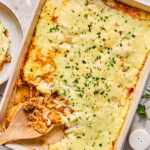 A baked casserole in a rectangular dish topped with melted cheese and chopped parsley, with one serving removed and a wooden spoon resting inside.