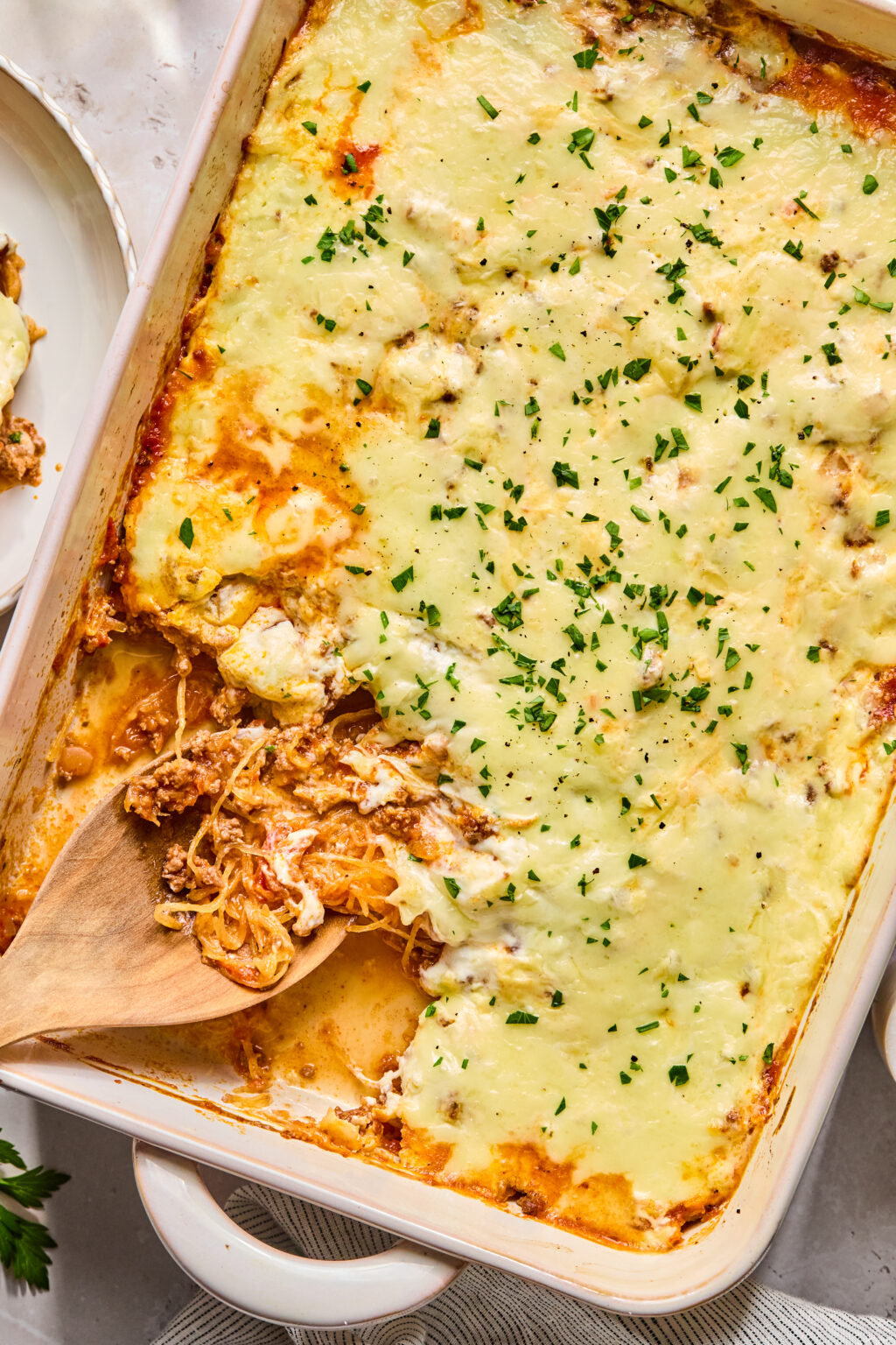 A baked casserole dish filled with cheesy spaghetti squash topped with chopped herbs, with a portion being served using a wooden spatula.
